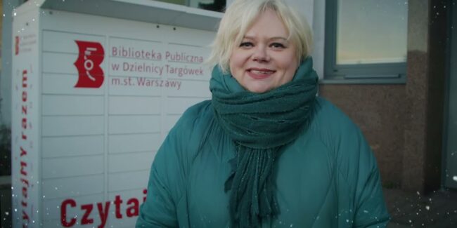 uśmiechnięta kierowniczka wypożyczalni nr 98 stoi przy książkomacie i poleca świąteczne nowości wydawnicze