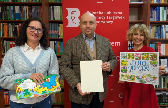 dyrekcja biblioteki i Koordynator Edukacji Kulturalnej w Dzielnicy Targówek pozują do zdjęcia razem z grami i książkami sensorycznymi, w tle regały wypełnione książkami