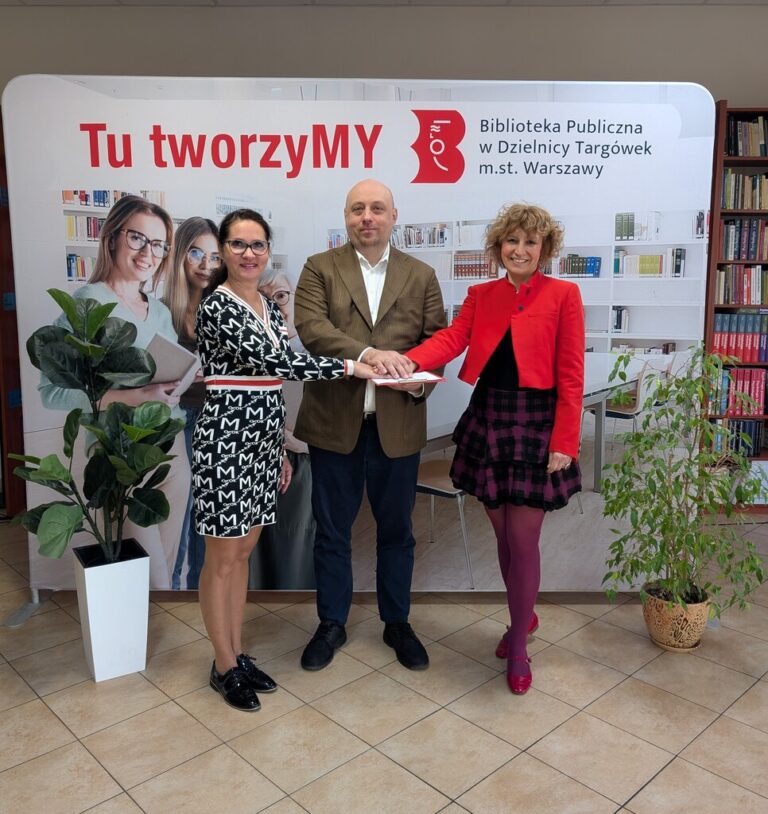 dyrekcja biblioteki i koordynatorka edukacji kulturalnej pozują do zdjęcia na tle ścianki z napisem Tu tworzymy, wokół nich kwiaty doniczkowe