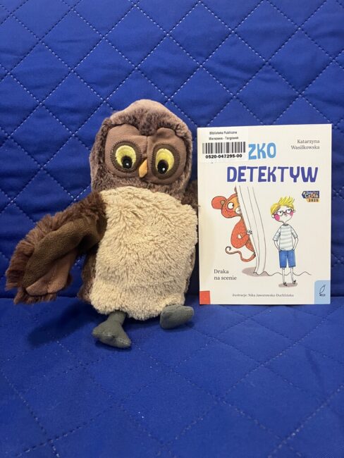 maskotka sowa Jadwiga trzyma skrzydłami polecaną książkę