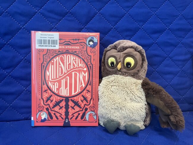 maskotka sowa Jadwiga trzyma skrzydłami polecaną książkę