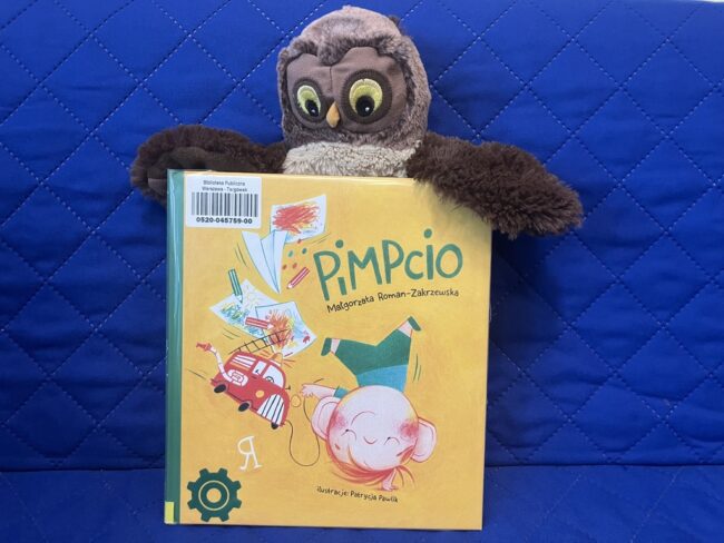 maskotka sowa Jadwiga trzyma skrzydłami polecaną książkę