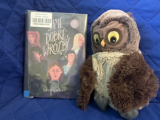 maskotka sowa Jadwiga trzyma skrzydłami polecaną książkę