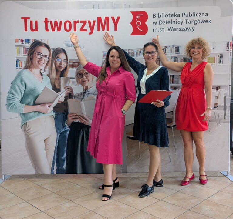 autorki projektu pozują do zdjęcia na tle ścianki ze zdjęciem biblioteki, logo biblioteki i napisem Tu tworzymy