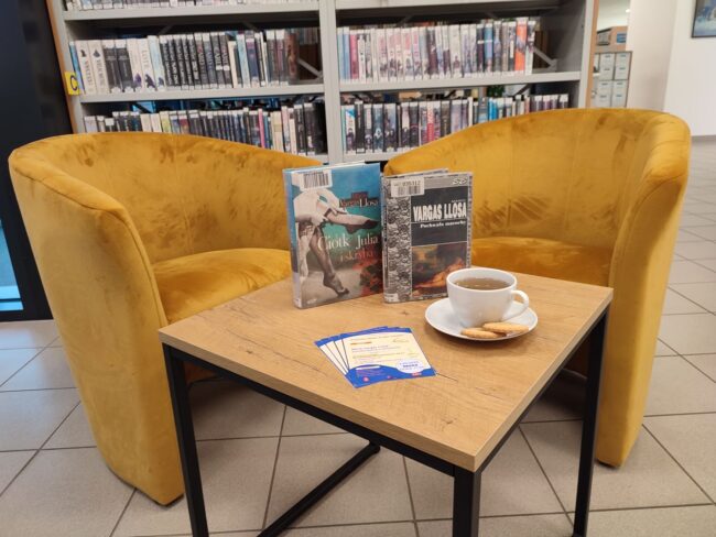 fotele, stolik z książkami omawianymi podczas spotkania, filiżanka herbaty i herbatniki, w tle regały wypełnione książkami