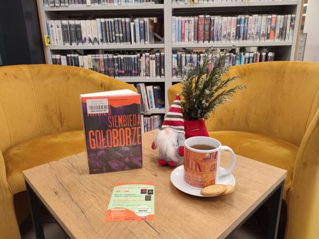 na stoliku w bibliotece jest książka, kubek z herbatą, w tle fotele i regały z książkami