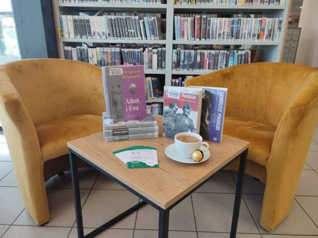 dwa fotele, stolik z książkami, filiżanka, ciastko, w tle regały biblioteczne