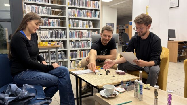 sesja RPG w bibliotece - 3 osoby przy stole grają w grę RPG